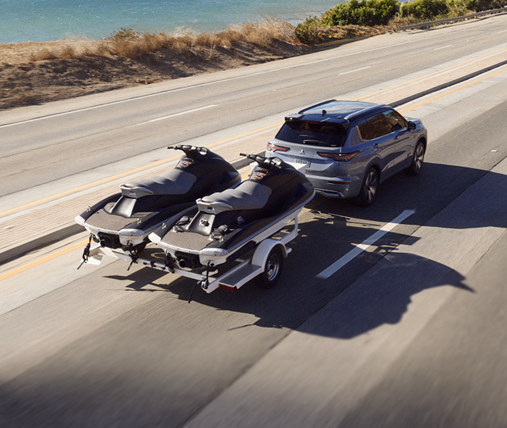 2026 Mitsubishi Outlander SUV driving on a highway beside water and towing two personal watercrafts while engaging the Trailer Stability Assist system.